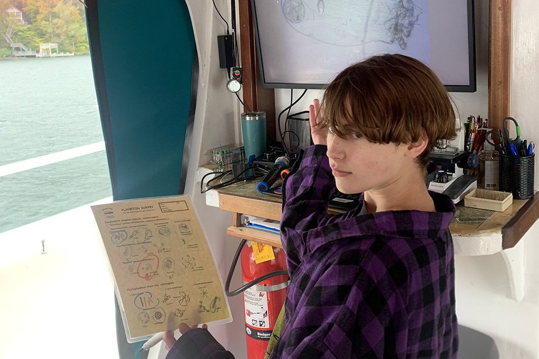 A child gestures toward a computer screen depicting bacteria while on a Floating Classroom cruise. Cayuga Lake is visible out the window as the student stands in the boat's interior.