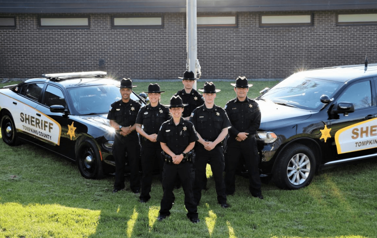 An outdoor photo in front of the County Police Department, 2 ploice cars together with a a group of 6 officers together.