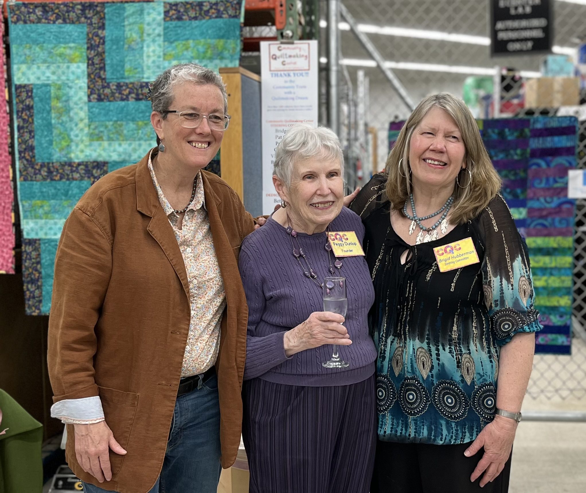 Diane, Peggy, and Brigid standing together in front of the display quilts