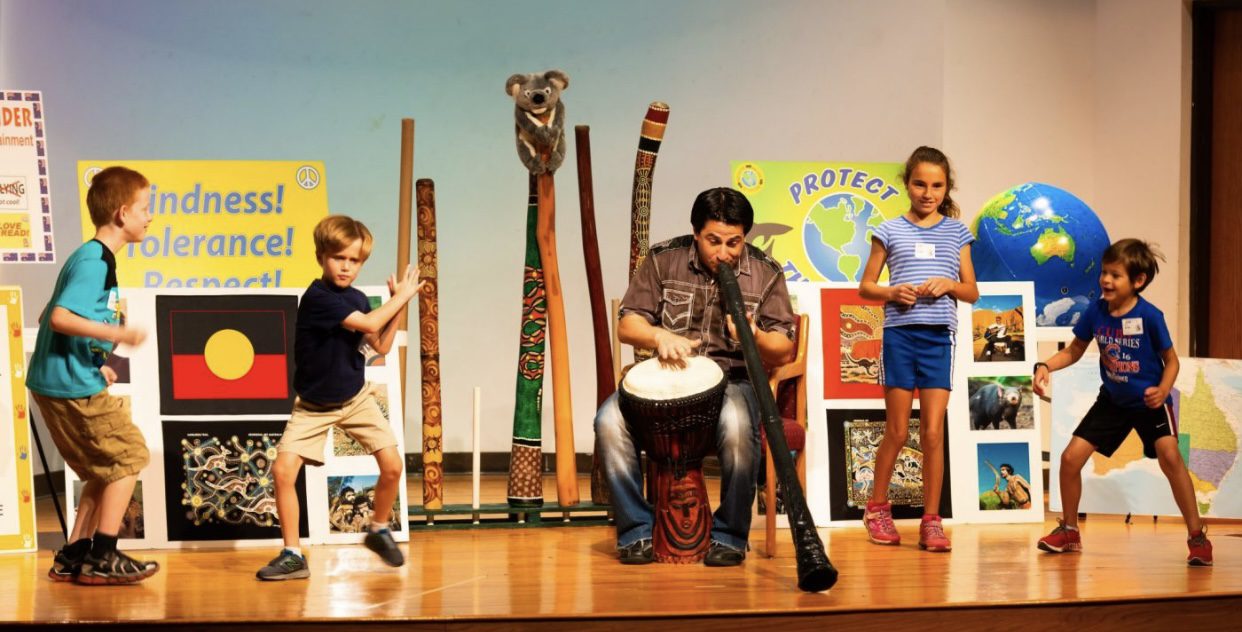Darren Liebman, from Didgeridoo Down Under giving a performance playing a drum and a black Didgeridoo on stage. More Didgeridoos are pictured behind him as well as many messages that Didgeridoo Down Under teaches in their performances.