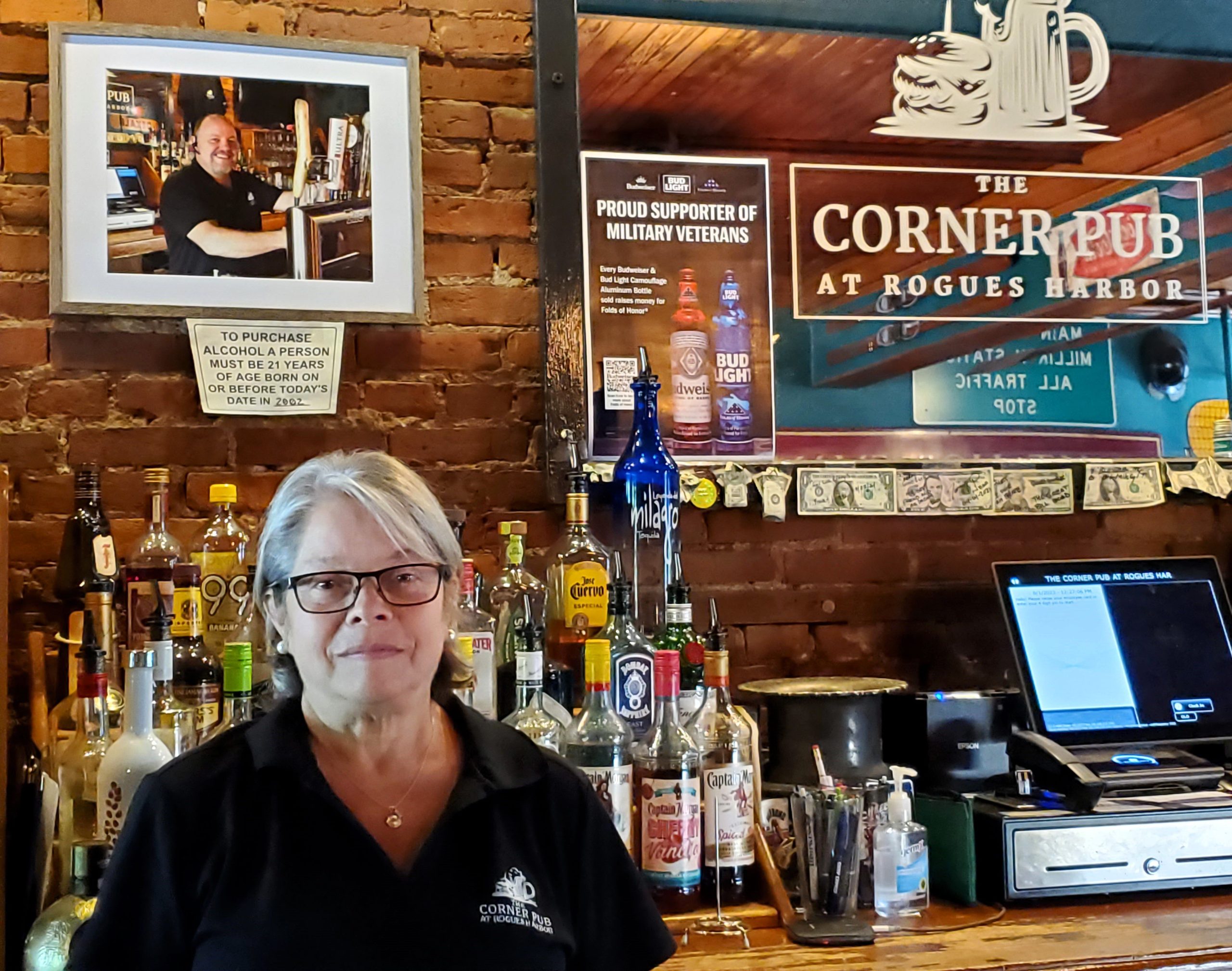 Kathy Leonardo stands behind the bar at The Corner Pub where a photo of Kerry Brace, her partner and founder of the Pub, looks on. This food-forward pub serves casual comfort food seven nights a week.