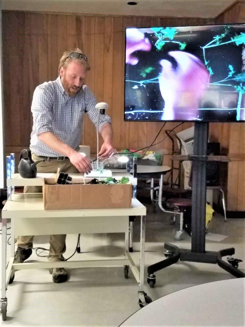 Dr. Jason Oliver, Agriculture Program teacher at Groton High School, demonstrating the correct way to transplant kale seedlings, with the help of his Elmo projector, during the Groton HARVEST Garden Plot Project class.