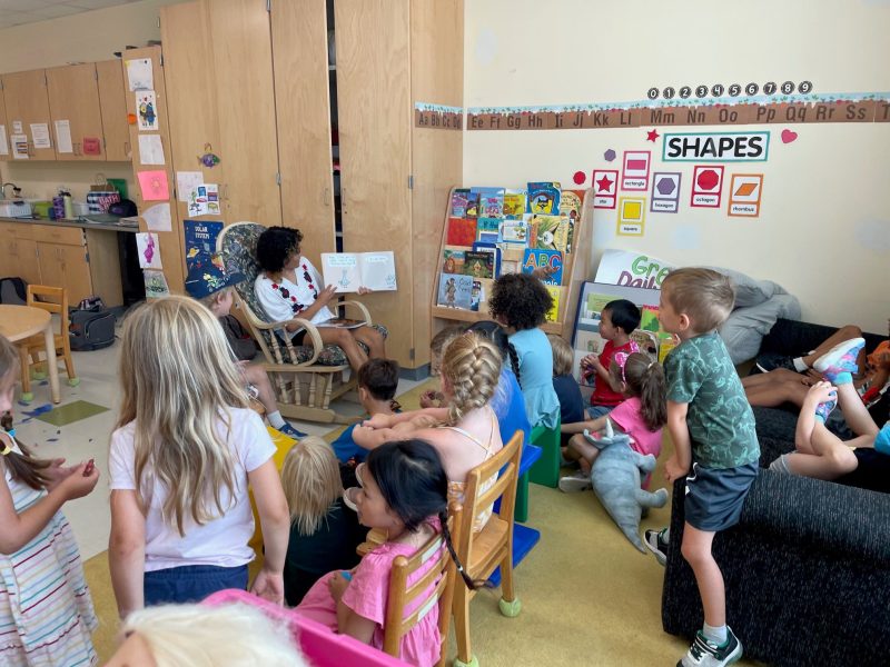 a group of pre-k students gather around a teacher/teaching assistant during story time