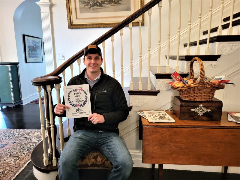 Drew Clare, vice president and general manager of The Benn Conger Inn, proudly displaying the inn's recent Top 3 Small Hotels in Tompkins, NY 2023 award from Travelmyth near the grandiose staircase in The Benn's foyer.