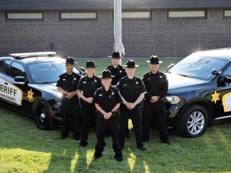An outdoor photo in front of the County Police Department, 2 ploice cars together with a a group of 6 officers together.