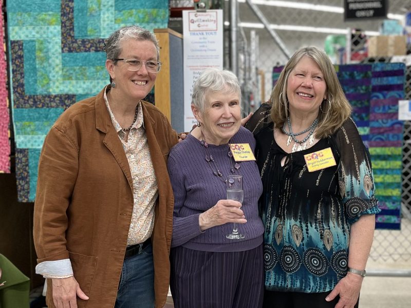 Diane, Peggy, and Brigid standing together in front of the display quilts