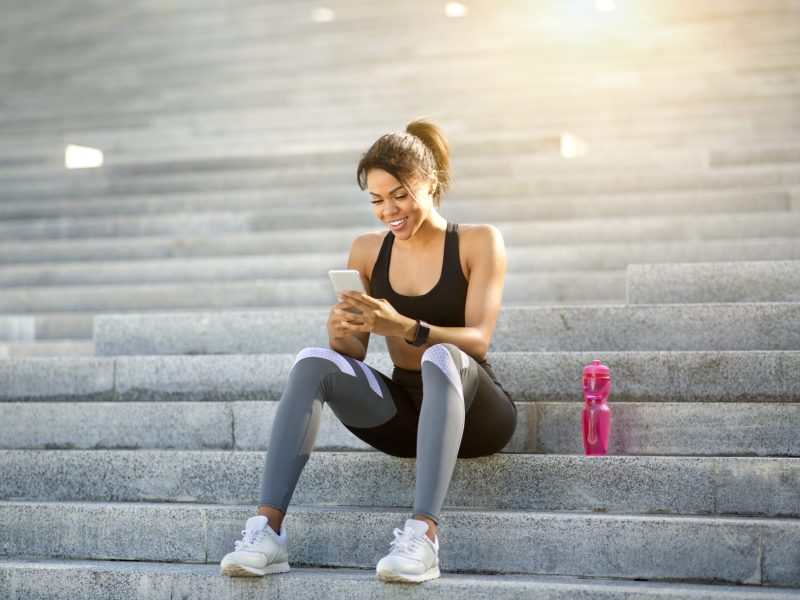 Sporty black girl using phone and drinking water while resting, training at park, copy space, sun glare