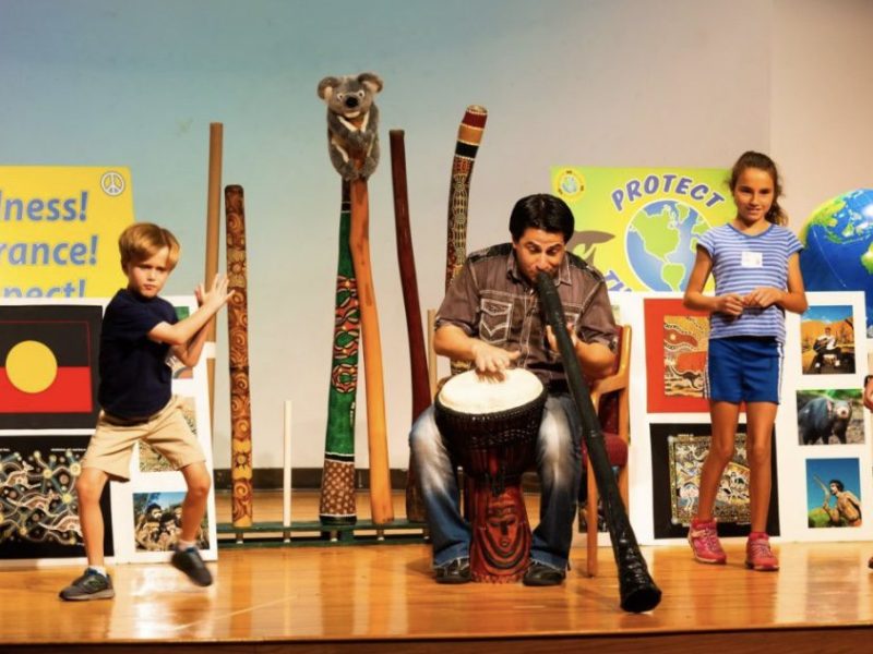 Darren Liebman, from Didgeridoo Down Under giving a performance playing a drum and a black Didgeridoo on stage. More Didgeridoos are pictured behind him as well as many messages that Didgeridoo Down Under teaches in their performances.
