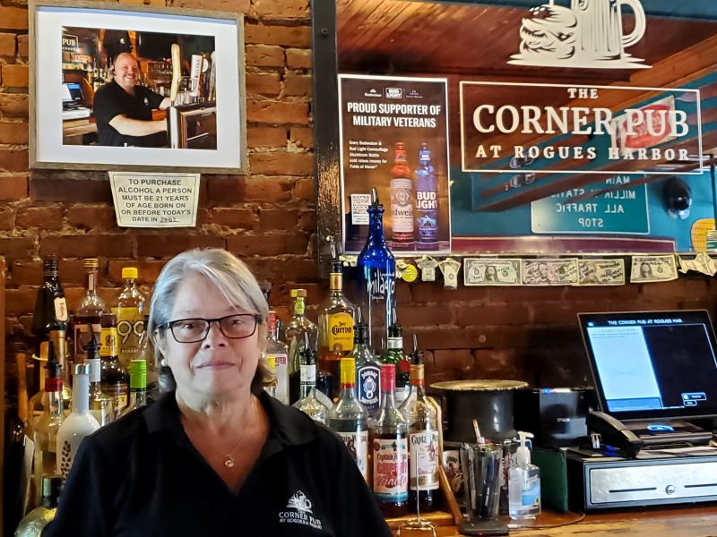 Kathy Leonardo stands behind the bar at The Corner Pub where a photo of Kerry Brace, her partner and founder of the Pub, looks on. This food-forward pub serves casual comfort food seven nights a week.